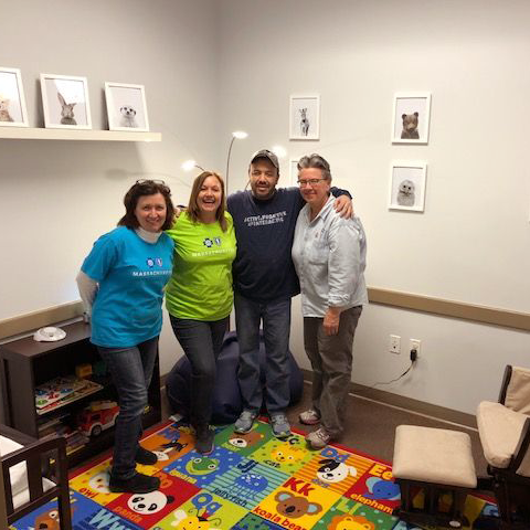4 adults stand with arms around each other smiling in a room with light walls and colorful floor covering.
